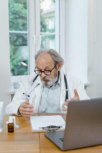 Elderly doctor explaining medication during an online consultation in a well-lit office setting.